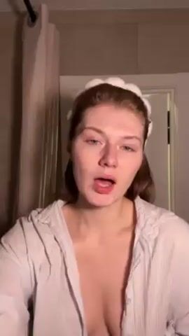 A young woman with light skin and auburn hair, wearing a white shirt and a white cloud-shaped headband, stands in a bathroom and touches her face and neck. She is looking at the camera, moving her mouth slightly. A shower curtain hangs to the left, and a door is visible in the background. Music plays in the background.