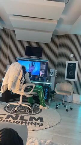 A person sits in a white office chair in front of a large computer monitor. The monitor displays a video showing hands and a lit candle. The person is wearing green pants, a white shirt, and white shoes. A white jacket hangs over the back of the chair. The room has a modern design with vertical wood panels on the wall, white acoustic panels on the ceiling, and a circular rug with the words "CNLM PUBLISHING" on the floor. There is a smaller monitor above the main one, as well as speakers, a small chair, and framed speakers visible in the background.