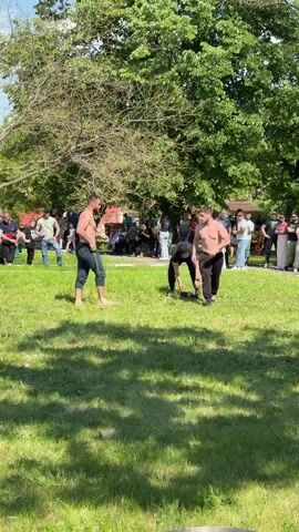 The video shows two bare-chested men wrestling on a grassy field surrounded by onlookers. The men are wearing dark-colored pants. Other men are wrestling nearby. Some people are sitting on benches while most are standing around the perimeter of the wrestling area. There are trees and buildings in the background. The sky is partly cloudy.