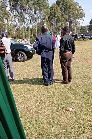 This media file depicts an outdoor scene, likely a funeral procession or a related event, in Lumakanda Ward, Kenya. The time appears to be daytime, under a clear, sunny sky. The setting is a grassy field bordered by a line of tall eucalyptus trees, suggesting a rural or semi-rural area.

The primary subjects are several men, most of whom are dressed formally. Two men in the foreground are facing away from the camera, one in a dark blue suit and the other in a black shirt and brown trousers. A third man in a brown suit is visible to the right. Other individuals, including one wearing a cap and a backpack, are also present, as are several vehicles.

Prominently featured is a black hearse with a white ribbon draped across its rear window. Another black SUV is parked nearby. Several flags, including one with red, white, and green stripes, are visible, possibly indicating a national or political affiliation. The overall atmosphere seems somber and respectful, consistent with a funeral gathering. There is a discarded bottle on the grass in the foreground. No discernable text is visible on the flags or vehicles.