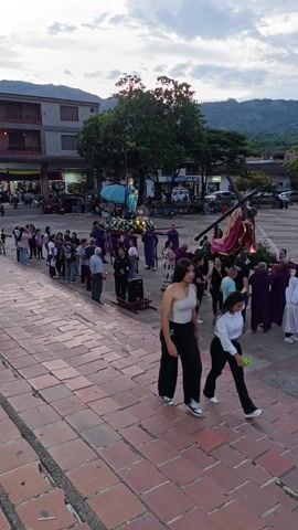 The video shows a religious procession taking place on a brick-paved plaza. A group of people in purple robes carry a figure of Jesus on a cross and another bearing an effigy of the Virgin Mary, both adorned with flowers and lights. Behind them, a line of onlookers observes the procession. A speaker on a stand amplifies the proceedings. Two young women walk in the foreground, while vehicles and buildings are visible in the background, set against a backdrop of mountains under a cloudy sky.