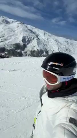 A snowboarder in a white jacket, black helmet, and orange-tinted goggles stands atop a snow-covered slope, the imposing, snow-capped peaks of a mountain rising in the background under a partly cloudy sky. The snowboarder gestures with their left hand towards the snowy terrain, revealing bright lime-green snowboard boots strapped to the board. The video appears to be shot from a first-person perspective, with the snowboarder as the focal point against the stark white landscape.