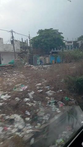 The video shows a neighborhood in a state of disrepair, seen from a moving vehicle. In the foreground, there is a significant amount of litter and discarded items covering the ground. Interspersed with the trash are patches of vegetation. In the background are small, modest buildings, some with visible signs of age and wear. Some appear to be residential while others could be small shops or businesses. The architecture is simple and functional. A few vehicles, including a truck and auto-rickshaws, are parked along the road. The sky is overcast and the overall light is muted. A person on a scooter is also visible.