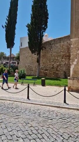 The video shows a cobblestone street with a chain fence running along the side. Behind the fence, there is a grassy area with two tall cypress trees. A stone wall forms the backdrop, with a white building visible on top. Two people, a man and a woman, are walking on the grass. The man is wearing a blue shirt and shorts, and the woman is wearing a white top and shorts. The sky is clear and blue. The video pans to the right, showing more of the stone wall and the white building, which appears to be an archway.