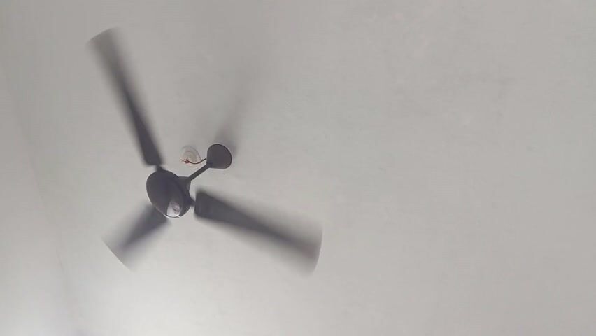 The video shows a dark-colored ceiling fan with three blades spinning rapidly against a white ceiling. The blades appear blurred due to the speed of rotation. The fan is centrally located in the frame but slightly offset to the left. A small white electrical component is visible near the fan's base. The ceiling has a slightly textured surface.