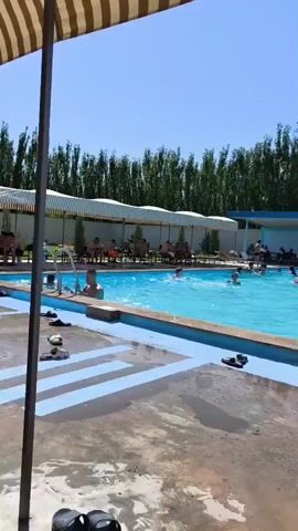 The video shows a crowded outdoor swimming pool on a sunny day. People are swimming, splashing, and relaxing around the pool area. There are lounge chairs set up under a striped awning, some of which are occupied. In the background, there is a building and trees. The water in the pool is bright blue, and the surrounding area appears to be concrete.