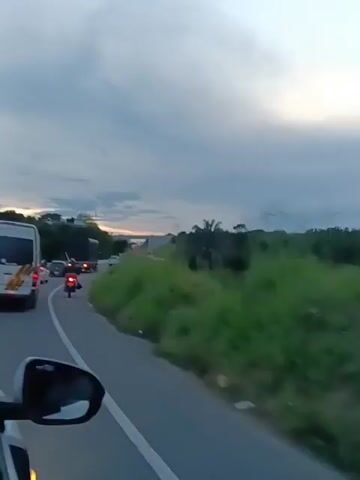 The video shows a road with cars and motorcycles driving on it. There is a grassy slope on the right side of the road. In the distance, a bridge can be seen under a cloudy sky. The video appears to be taken from a moving vehicle, as the perspective shifts. The lighting suggests it is either early morning or late afternoon.