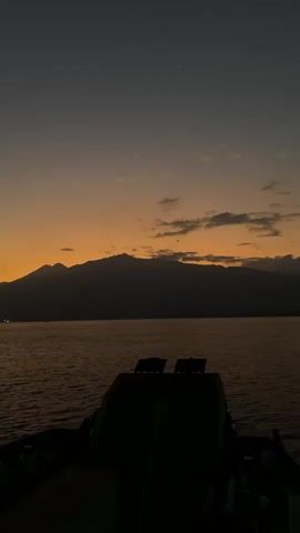 The video shows a sunset scene over a body of water. The sky is a gradient of dark gray to orange, with some clouds. In the distance, silhouetted mountains are visible. In the foreground, the deck of a boat is visible, partially in shadow. The water is dark and calm, with a few distant lights visible on the horizon. The overall mood is peaceful and serene.
