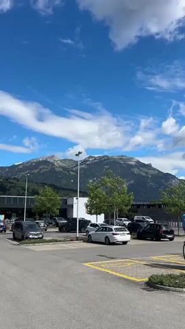 The video begins by panning across a parking lot with several cars parked, a large building in the background, and mountains under a blue sky with scattered clouds. In the foreground, there is a silver convertible with its top down. As the video continues to pan, more cars and trucks are visible in the parking lot. The building in the background appears to be a commercial establishment with people walking in front of it. The camera continues to move, showing more of the parking lot and the surrounding mountainous landscape. Towards the end of the video, the camera focuses on a yellow car parked in the lot.