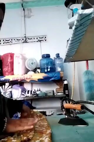 A person is shown ironing a piece of fabric. The fabric is yellow with a floral pattern in various colors. The person is wearing a brown garment, possibly a hijab or abaya. They are holding an iron with an orange handle and are pressing it down on the fabric. The ironing board is blue. In the background, there are shelves with various items, including plastic water bottles, a fan, and other objects that are difficult to identify. The room appears to be a storage area or workshop.