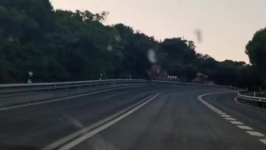 The video shows a perspective from a moving vehicle on a winding road. The road has a guardrail on the right side and a double white line marking the center. Trees and vegetation line the sides of the road, with some power lines visible in the background. There are traffic signs, including a sign indicating a sharp curve and a speed limit of 40. The sky is overcast, suggesting it might be dawn or dusk. There are no people visible in the video.