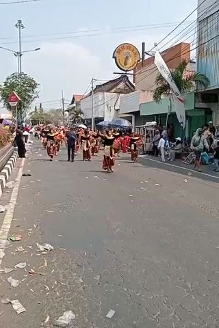 The video shows a street parade with several people in traditional costumes dancing. They are moving down a paved road lined with buildings and spectators. The dancers are wearing colorful skirts, tops, and headpieces. Some of the dancers are carrying baskets. The spectators are watching the parade from the side of the road. The weather appears to be sunny.