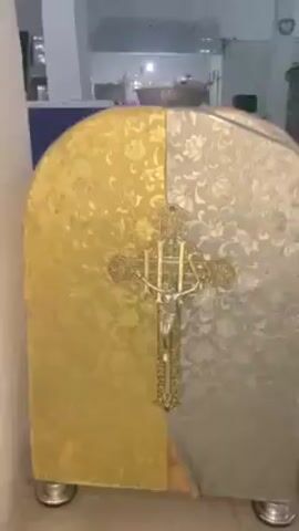 The video shows a cabinet with a crucifix on its front. The cabinet is divided into two halves: one is gold, the other silver. Both halves have a floral pattern. The crucifix is silver in color. The cabinet is on short, silver legs and is in a room with white walls and tile floors. In the background, there is a kitchen area.