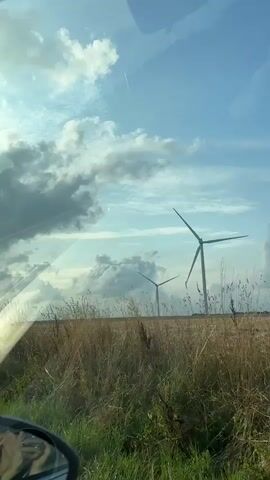 This is a vertical video taken from inside a vehicle, looking out through the windshield. The scene depicts a rural landscape with a field of dry grass or wheat in the foreground, bordered by a metal guardrail. Two large wind turbines are visible in the middle ground, with their blades in motion. The sky is blue with scattered white and gray clouds. The image is slightly distorted by reflections on the windshield.
