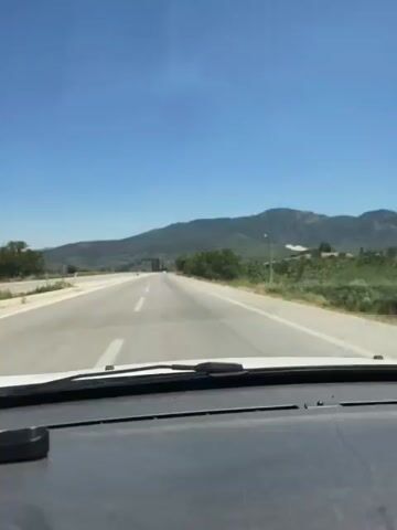 This video shows a first-person view from inside a car driving on a highway. The road is paved and has white lines marking the lanes. On either side of the road are grassy shoulders and some trees and hills in the distance. The sky is blue and clear. The camera is positioned to capture the view through the windshield. There is a dashboard visible at the bottom of the frame. The video appears to be taken on a sunny day.