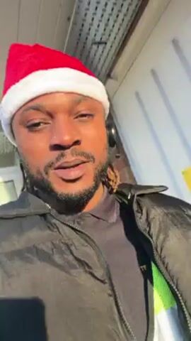 A man wearing a red Santa hat, black jacket, and a neon yellow safety vest with a black shirt underneath, is standing outside in front of a white wall with a "Gas Meter" sign. He has a nose piercing, a beard, and dreadlocks. He is wearing headphones and talking to the camera, wishing viewers a Merry Christmas. Visible breath indicates a cold outdoor temperature.