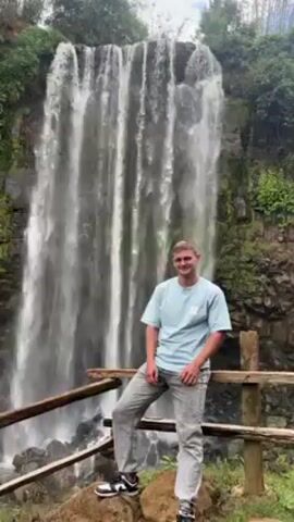A man is standing in front of a large waterfall, leaning against a wooden railing. He is wearing a light blue shirt, light grey pants and black and white shoes. A woman approaches and he scoops her up in his arms, and they share a kiss. The woman is wearing a white top and light beige pants and white shoes. The waterfall is surrounded by green foliage.