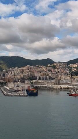 The video shows a port city scene with a variety of boats, buildings, and ships along the harbor. The camera is oriented vertically, showing the city rising up the hill. The sky is partially cloudy, with a mix of blue sky and grey clouds. A ship’s masts are briefly visible in the upper right of the video.