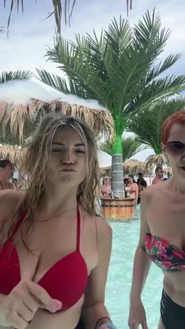 This video shows two women in swimsuits posing in a pool at a beach club. Palm trees and thatched umbrellas are visible in the background. Other people can be seen in the water and at a bar. The weather appears to be sunny and warm, suggesting a daytime event. The women are smiling and appear to be enjoying themselves, possibly taking selfies or recording a short video for social media. The scene evokes a tropical vacation or a day at a summer resort. No visible text is present in the video.