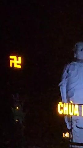 The video shows a nighttime scene. On the left side, there's an illuminated swastika symbol. Next to it is a slightly blurred smaller object emitting dim lights. On the right, a statue stands partially lit, with only its upper half visible. The statue's features are indistinct due to the lighting. Underneath the statue, the words "CHÙA B..." are illuminated, possibly a sign indicating a temple. The background is dark, indicating a nighttime or low-light environment.