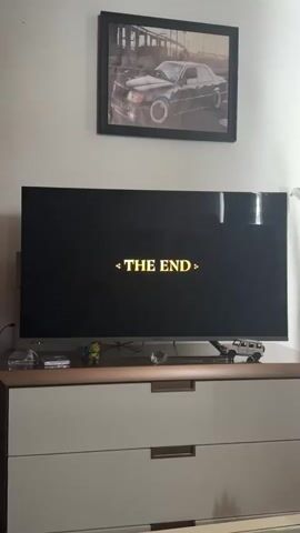 The video shows a living room space. The central focus is a modern television displaying the words "THE END" in a golden, ornate font. Below the TV is a low dresser with two drawers, brown on top and white drawers with rectangular metal drawer pulls. On the dresser, besides the TV, there are various small items, including what appears to be an ashtray, a small Ninja Turtle figurine, a small crystal paperweight, and a white toy car. Above the TV, a framed picture of a black Mercedes-Benz hangs on the white wall. As the video progresses, the screen goes black and the sounds of movie credits begin to fade.