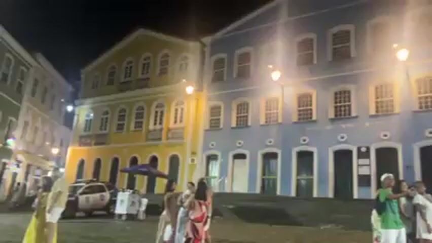 At night, in what appears to be a public square, there are several brightly colored buildings. The building on the left is yellow, the building in the center is blue, and buildings on the right are primarily white with yellow accents. The blue building has a banner that reads “QUANTA FREE” hanging on it. Several people are milling about in the square, including some posing for pictures in front of the blue building. A small service vehicle is parked to the left of the yellow building. Street lamps illuminate the square.