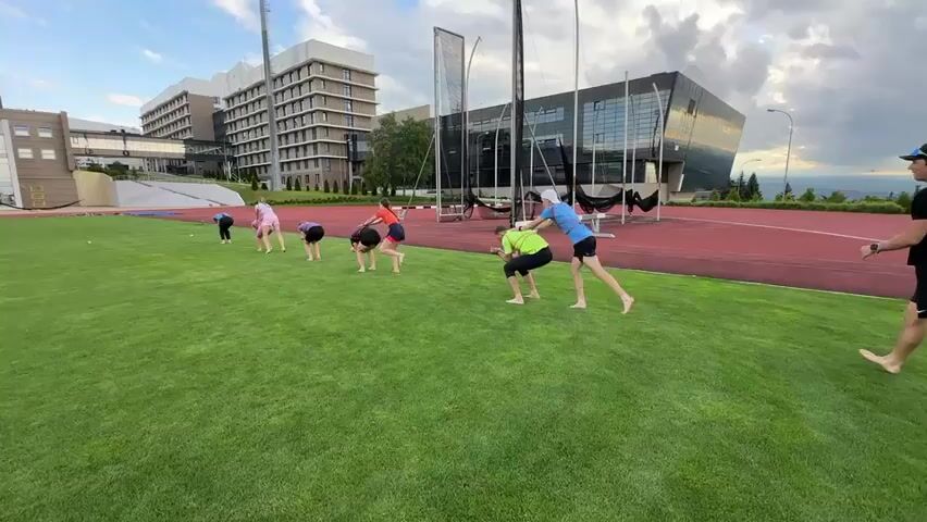 A group of nine people, mostly young adults, are positioned on a vibrant green grassy field in front of a red running track. They appear to be engaged in some form of outdoor training or exercise. Seven individuals are lined up in a row on the grass, bent over with their hands touching the ground, as if preparing for a race or an agility drill. Two individuals stand upright further to the right on the grass, observing the group. One of these standing individuals, wearing a bright green t-shirt and black shorts, is in a crouched position, possibly demonstrating an exercise or giving instructions. The other standing individual, wearing a black t-shirt and shorts, is looking towards the group. In the background, there are modern buildings with large windows, a tall stadium light, and some netting or large equipment, suggesting a sports facility or university campus. The sky is partly cloudy, with a mix of blue sky and white clouds. The overall scene suggests an active and organized event taking place outdoors.