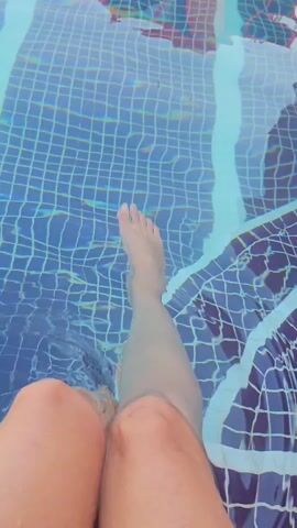 A first-person perspective video shows legs submerged in a swimming pool. The legs are visible from the knees down. The pool water is clear and blue, with a tiled bottom. The tiles appear to be blue and white in a grid pattern. The person's legs are moving slightly, creating ripples in the water. The video is taken from a slightly downward angle.