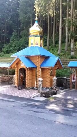 The video shows a small, wooden Orthodox chapel with a blue roof and a golden dome adorned with a cross. The chapel is situated in a forested area with tall trees and lush greenery, suggesting a natural and peaceful setting. In front of the chapel, there is a paved area with decorative tiles, and to the right, a wooden sign displays religious icons. The overall scene evokes a sense of tranquility and reverence, possibly a place of worship or a pilgrimage site in a rural Ukrainian landscape. The time of day appears to be daytime, with sunlight filtering through the trees, indicating fair weather. There are no people visible in the video. The location context provided specifies Synevyrska Poliana, Ukraine, which aligns with the visual cues of a mountainous, forested region often associated with such religious structures in Eastern Europe.