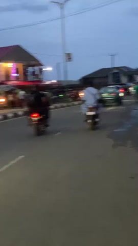 The video shows a street scene at dusk. The sky is a muted blue-gray, suggesting the end of daylight. In the foreground, the street is wet in places, reflecting the ambient light. A car with illuminated red tail lights drives away from the camera. Two motorcycles, each with a single rider, are seen driving in the same direction as the car. Several people are walking and standing along the side of the road. Buildings and streetlights are visible in the background, with a sign mounted on a pole. The scene has a casual, everyday feel, suggesting a typical evening in a town or village.
