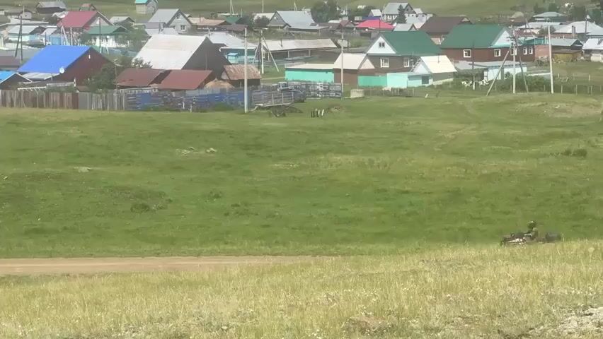 A person on a dark-colored ATV drives down a dirt road that cuts through a grassy field. In the background, a village of houses with colorful roofs and wooden fences is visible. The sky is overcast. The ATV is positioned in the center of the frame, moving from left to right. The surrounding landscape is predominantly green, with tall grass in the foreground.