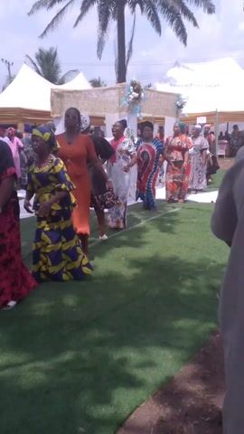 The video shows a group of women in colorful, patterned dresses walking on green turf. They are near white tents and a decorated archway, under the shade of a palm tree. Some of the women are wearing headwraps. One woman pauses in the line to sing into a small microphone.
