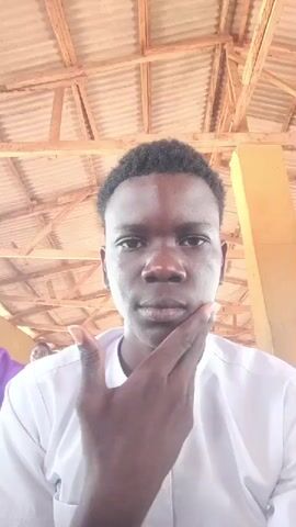 This is a short video clip of a young Black male filmed from the chest up, in a close-up shot. He is wearing a white traditional garment with a high collar and intricate button detailing. His skin is dark, and his hair is short and curly. The man is looking directly at the camera and makes a series of facial expressions, touching his face and chin with his fingers. He appears to be indoors, possibly in a sheltered outdoor space, as indicated by the wooden beams and corrugated metal roof visible above him. The lighting suggests it is daytime. In the background, two other individuals are partially visible, suggesting a public or social gathering. The overall mood is casual and personal, as the subject interacts directly with the camera. There is no visible text in the media file.