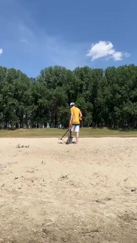 A person wearing a yellow t-shirt, white cap, and shorts is metal detecting on a sandy area. They are holding a metal detector in their right hand and a shovel in their left. The scene is outdoors, with a line of green trees in the background and a blue sky with scattered white clouds above. The ground is dry and sandy, with some sparse vegetation. The sun appears to be high in the sky, suggesting it is daytime. The overall atmosphere seems peaceful and recreational, with the person engaged in a hobby. There is no visible text in the frame.