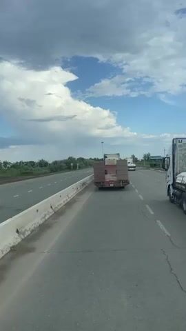 The video is shot from the perspective of a vehicle driving on a highway. A truck with a flatbed trailer carrying what appears to be boxes is in front of the camera vehicle. Another truck is visible in the distance on the right side of the road. The highway is bordered by a concrete barrier on the left and a grassy shoulder with some trees on the right. The sky is partly cloudy with patches of blue sky. The road surface shows signs of wear and tear, with visible cracks.