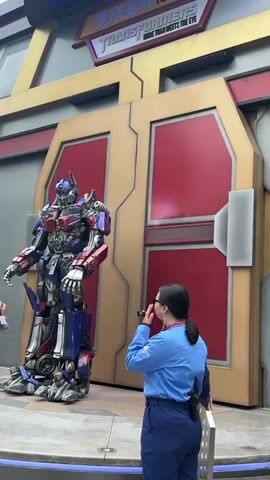 The video opens with a medium shot of a large, detailed Optimus Prime costume standing on a platform in front of a large, golden-brown and red door. The Optimus Prime costume is blue, red, and silver, with intricate mechanical details. A person wearing a blue uniform and dark pants stands to the right of the Optimus Prime costume, facing away from the camera and towards the robot. They appear to be a staff member, possibly guiding guests or controlling the animatronic. The background features the "TRANSFORMERS" logo, suggesting the location is a theme park or attraction. In the later part of the video, another person wearing a dark shirt and shorts enters the frame from the left and approaches Optimus Prime. They interact briefly with the robot, possibly posing for a photo. The lighting is bright and appears to be natural daylight. The scene is static, with no significant movement from the robot.