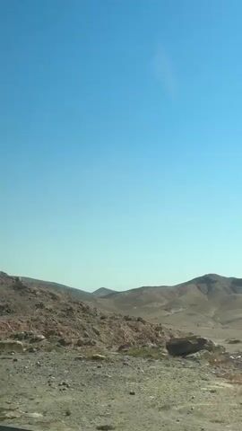 The video shows a desolate, arid landscape with rolling hills under a clear, bright blue sky. The terrain is rocky and sandy, with sparse vegetation. The camera is likely positioned inside a moving vehicle, as the landscape scrolls by. The overall impression is of a dry, deserted region, possibly a desert or semi-desert environment.