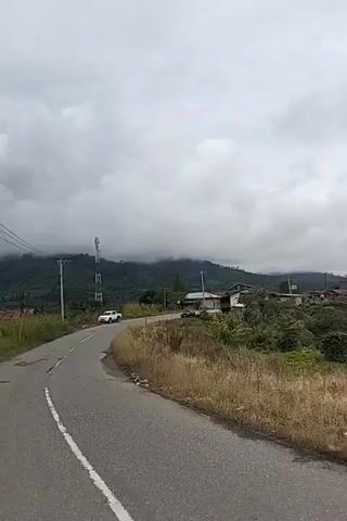 The video shows a person filming from a moving vehicle on a paved road. The road curves to the left and passes by a grassy embankment on the right. In the background, there are several houses and a cell tower. The sky is cloudy and the landscape appears mountainous and green. The video is short, lasting only a few seconds, and the camera is pointed slightly downward, capturing the road ahead and the surrounding environment. There are a few vehicles visible on the road, including a white SUV and a dark sedan. The houses appear to be situated on a hillside overlooking the road. The overall impression is of a rural or semi-rural setting with overcast weather.