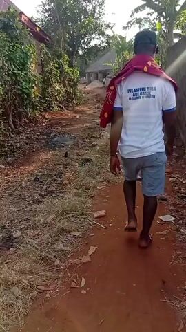 A man is walking away from the camera down a dirt road in what appears to be a rural area. The road is lined with vegetation on one side and a brick wall on the other. The man is wearing a white T-shirt, blue shorts, and a dark baseball cap. He has a red cloth draped over his shoulder. There are houses visible in the distance. The sky is bright and the sun appears to be shining. The camera seems to be held by someone walking behind the man, and their finger partially obscures the view at times. There is some trash on the ground along the road.