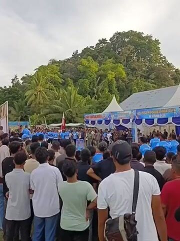 The video depicts an outdoor gathering of a large crowd of people, likely a community event or rally, at Keude Trienggadeng, Indonesia. The subjects are predominantly young men, many dressed in casual attire. Some are wearing light-colored t-shirts, while others are in darker clothing. A significant number of people in the front rows are facing away from the camera, looking towards a stage or a central point of activity. In the background, there is a tent with blue and white banners, suggesting an organized event. Behind the tent, a lush, green, tree-covered hill rises, indicating a tropical or subtropical environment. Palm trees are also visible, further supporting this assumption.

The activity taking place appears to be a speech or presentation, as several individuals are standing on a raised platform or near the tent, some of whom seem to be speaking. The overall atmosphere seems to be one of communal gathering and engagement, though specific emotions are difficult to discern from this distance. The time of day appears to be late afternoon or early evening, as the light is soft and the sky is a pale blue, with some hints of clouds.

Visible text on the banners includes "HARI BHAKTI ADHYAKSA" and "PADA KEPOLISIAN PIDIE JAYA". There is also text that appears to read "KE-X TAHUN 2026", suggesting the event is related to the Public Prosecutor's Office (Kejaksaan) and is in commemoration of its anniversary in 2026. The presence of police uniforms on some individuals on the stage might indicate a collaboration or a joint event. The scene is set on what appears to be an open field or sports ground, with a basketball hoop visible on the left side of the frame.