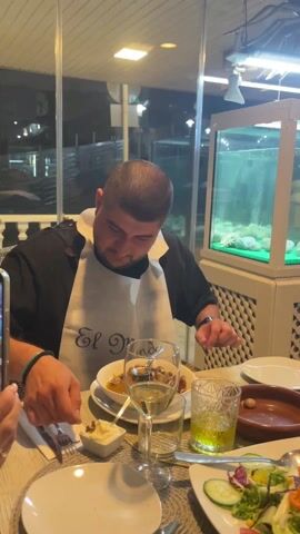 The video shows a man in a restaurant setting, dressed in a black shirt and wearing a white bib with the inscription "El Mundo". He is seated at a table laden with food and drinks. The table features a bowl of food in front of him, a glass of white wine, a small dish of what appears to be aioli, a glass filled with a yellow liquid (possibly olive oil), a plate with salad, and an empty plate. To his left, someone is holding a smartphone, possibly recording. The man uses a small tool, possibly for extracting food, from the bowl and pauses. Then someone points in the air towards him and speaks, and he looks up and to the right. Behind him is a glass wall looking out to a dark outdoor area, and beside him is a decorative fish tank with what appears to be clam shells inside.