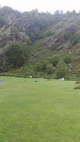A wide shot of a scenic landscape with a waterfall cascading down a rocky, tree-covered mountainside. In the foreground, a neatly manicured green lawn is dotted with picnic tables and some white signs. A paved path curves through the lawn, leading towards the base of the waterfall. The sky is overcast, suggesting a possibly cool or damp day. The overall impression is one of natural beauty and a recreational area.