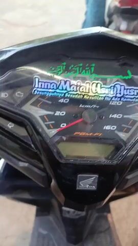 The video shows a person on a motorcycle at a gas station. The motorcycle dashboard is visible in the foreground. In the background, there are two people in red uniforms, likely gas station attendants, interacting with another person standing next to a motorcycle. There are also bamboo plants and a gas pump visible in the environment. The license plate on the motorcycle in the background reads "M 5086 AC".