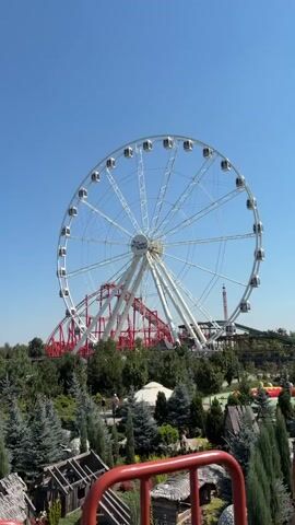 The video displays an amusement park scene under a clear blue sky. A large white Ferris wheel dominates the background, with a red roller coaster track visible behind it. In the foreground, there are miniature buildings, green trees, and red railings. The camera appears to be positioned on a ride, looking out over the park. There are no people clearly visible in the frame. The overall atmosphere is bright and sunny.