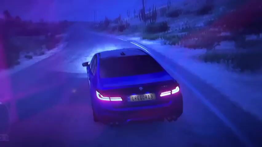 A blue car with glowing tail lights drives down a desert road at night. The car's headlights illuminate the road ahead, and the surrounding desert landscape is visible on either side. The sky is dark and appears to have some purple and pink hues. The license plate on the car reads GREGOR13.