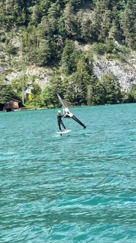 A person wearing a black wetsuit and a yellow helmet is wing foiling on a turquoise body of water. They are standing on a white surfboard with a hydrofoil attached, which lifts the board out of the water. The person is holding a black and white wing, using it to catch the wind. In the background, a steep, tree-covered mountainside rises from the water. A small wooden building is visible on the shore. The water is calm with small ripples. The sky is clear and bright.
