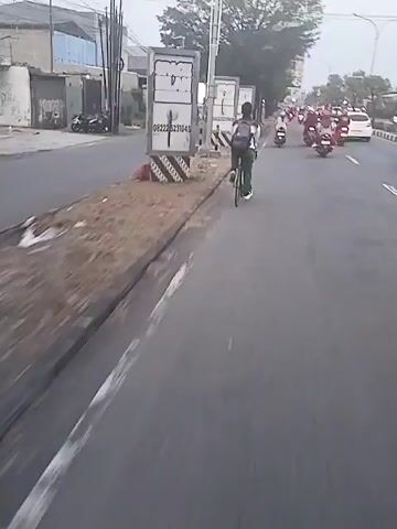 The video shows a person riding a bicycle on a street. The person is wearing a white and black jacket, green pants, and a backpack. They are riding on the left side of the road, next to a curb and some metal containers. There are other vehicles on the road, including motorcycles and cars. The surroundings appear to be urban or suburban. The camera seems to be mounted on a moving vehicle, likely another bicycle or motorcycle. The weather appears overcast.
