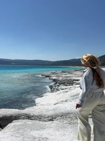A person with blonde hair is walking on a rocky, white shore next to a bright blue lake under a clear sky. The person is wearing a grey hooded jacket and beige pants. They are walking away from the camera towards the lake. The landscape is rocky and barren with mountains in the distance. The video shows the person taking steps forward with their right foot.