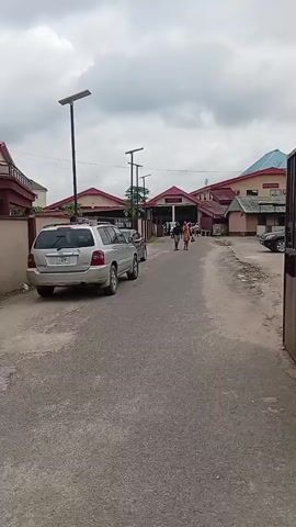 The video shows an outdoor scene with several buildings and a paved road. In the foreground, a silver SUV is parked on the left side of the road. Further down the road, two people are walking away from the camera. To the right, a large metal gate is partially open, revealing a glimpse of a car parked behind a wire fence. In the background, there are more buildings, some with red roofs, and others with green accents. The sky is overcast with clouds. The video pans and moves, offering different perspectives of the environment, including a shot of a sign with text that includes "BIBLE" and "BASIC INSTRUCTION BEFORE LEAVING THE EARTH." Another sign with directional arrows is visible near the gate. There are also solar-powered streetlights along the road. The scene appears to be a campus or a complex with multiple structures and an open area.