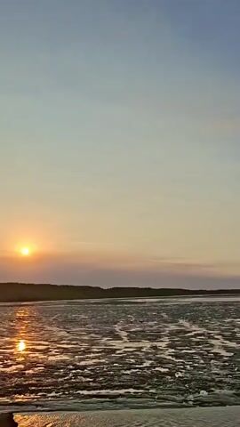 The video shows a wide, flat expanse of mudflats under a pale blue sky. The mudflats appear to be partially submerged in water, creating a textured surface with ripples and channels. In the distance, a few poles with red and white markers are visible, likely navigational aids. The lighting suggests it is either sunrise or sunset, with a soft glow on the horizon. The camera is moving, panning across the landscape, giving the viewer a sense of walking or driving through the scene. No people are clearly visible in the frame.